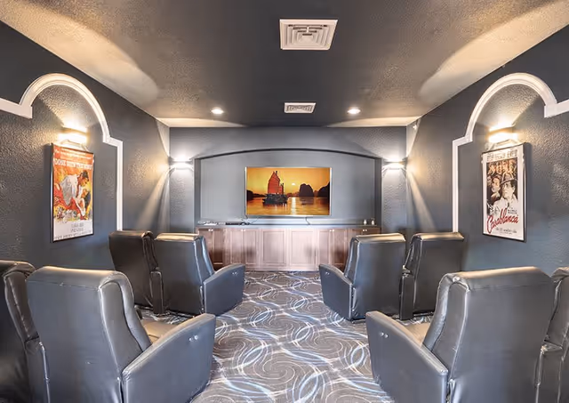 A small theater room with six black leather recliner chairs arranged in two rows facing a flat-screen TV mounted on a dark gray wall. The room has dark walls with white trim, two wall sconces providing light, and framed movie posters on each side wall. The carpet has a swirling pattern in shades of gray and blue.