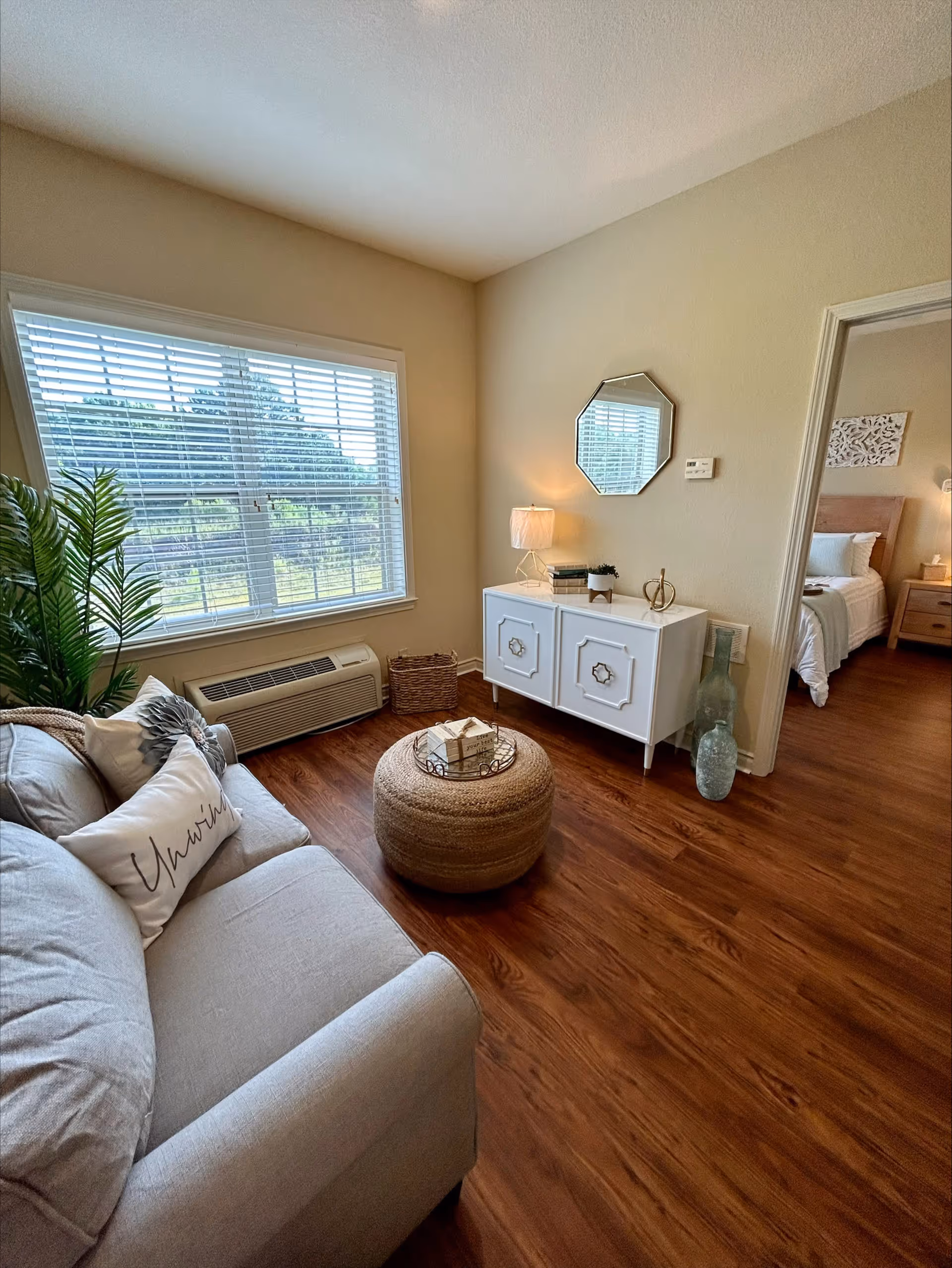 A cozy living room with a light gray sofa adorned with decorative pillows, a round woven ottoman with books on top, a white cabinet with a lamp, books, and decorative items, a large window with white blinds, and a view into a bedroom with a neatly made bed and nightstand.