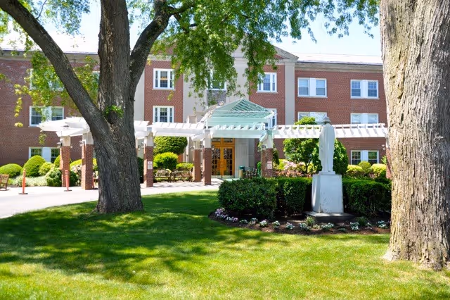 Exterior view of a three-story brick building with white-framed windows, a covered entrance with a white pergola, and a statue on a pedestal in a landscaped garden with green grass and trees.