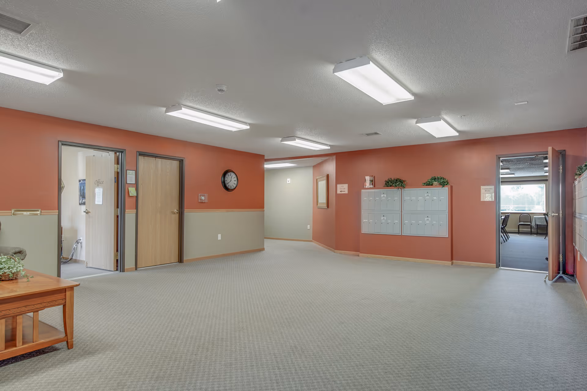 Interior hallway area of Keller Lake Commons with beige carpet and walls painted in two tones of red and beige. There are multiple doors, a wall clock, a set of mailboxes mounted on the wall, and fluorescent ceiling lights. A wooden table with a plant is partially visible on the left side.
