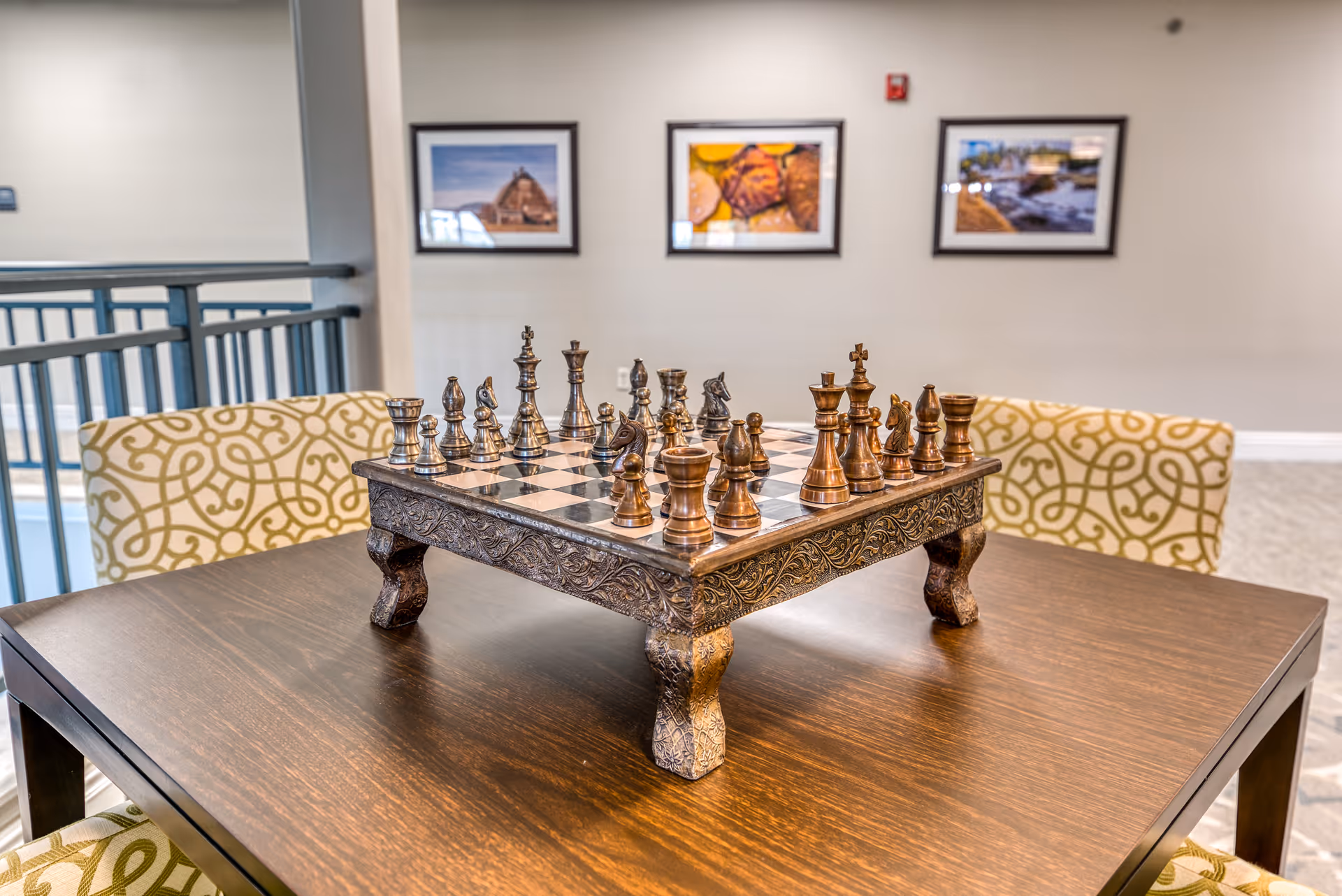 A decorative chess set with intricately carved wooden pieces on a raised chessboard placed on a wooden table. The table is surrounded by chairs with patterned upholstery. In the background, there are three framed pictures hanging on a beige wall.