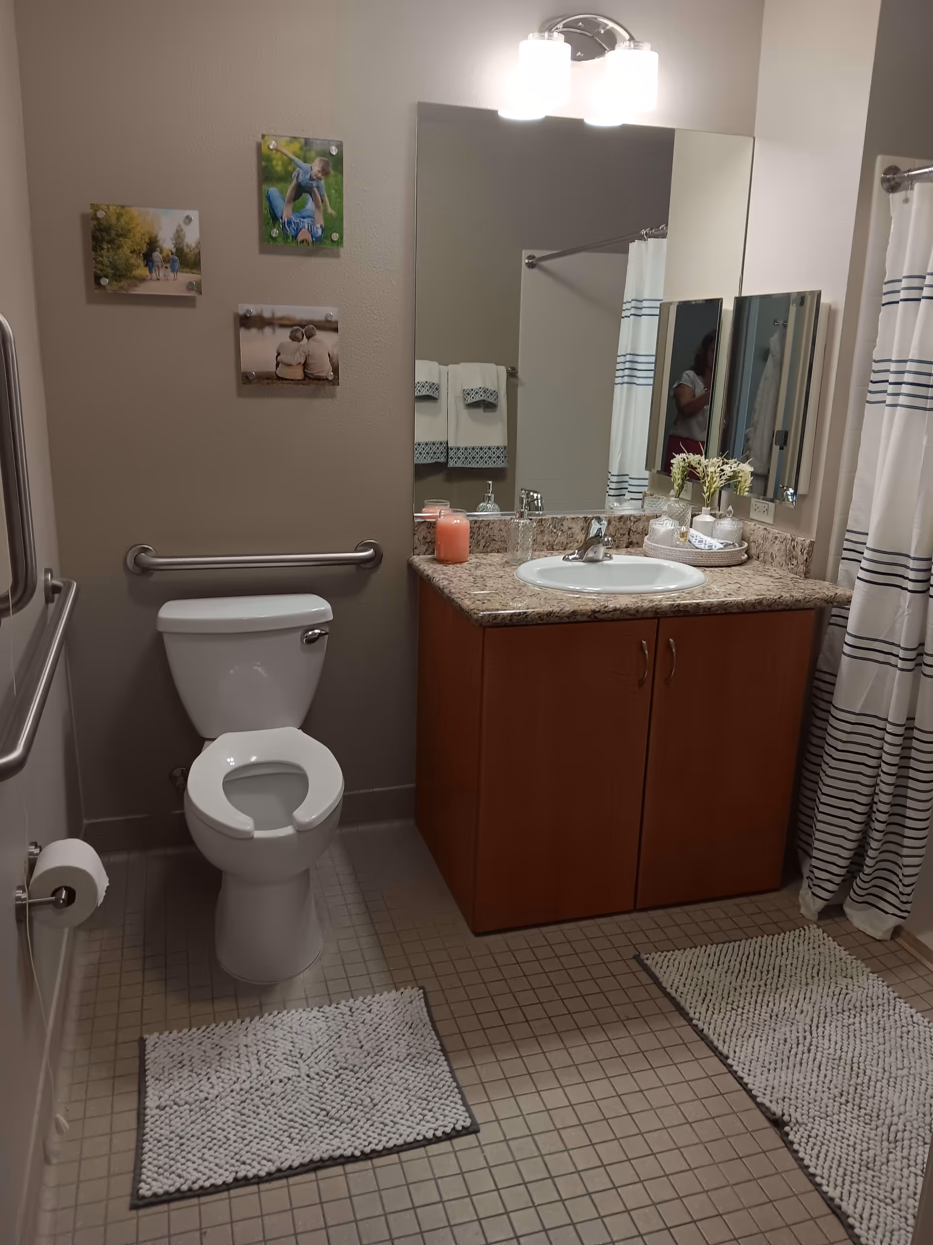 A clean bathroom with a white toilet, a wooden vanity with a granite countertop and a sink, a large mirror above the sink, and a shower with a white curtain featuring blue stripes. There are three small framed pictures on the wall above the toilet, two white textured bath mats on the tiled floor, and a wall-mounted light fixture above the mirror. A grab bar is installed next to the toilet, and towels are hanging on a rack reflected in the mirror.