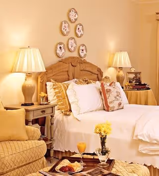 A cozy bedroom with a wooden headboard bed adorned with multiple pillows and white bedding. Two bedside tables each hold a lamp with a beige shade. Above the bed, there are six decorative plates arranged on the wall. In the foreground, there is a cushioned armchair and a small table with a vase of yellow flowers and a tray with food and drink.