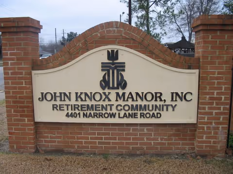 A large brick sign with a cream-colored panel displaying the text 'John Knox Manor, Inc Retirement Community 4401 Narrow Lane Road' with a decorative emblem above the text. The sign is outdoors with grass in front and trees and a gazebo visible in the background.