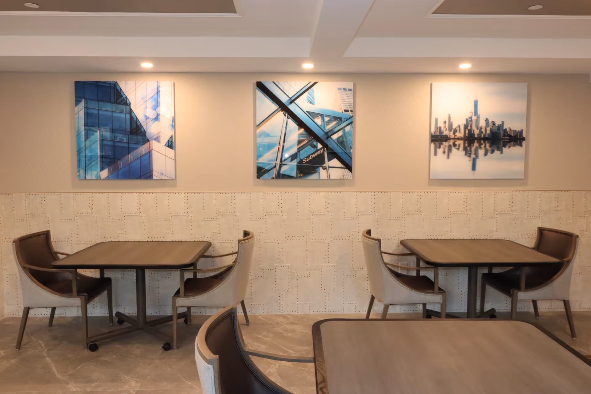 A dining area with two square tables, each surrounded by four cushioned chairs with armrests. The walls are decorated with three framed pictures featuring modern architectural and cityscape images. The floor is tiled and the ceiling has recessed lighting.