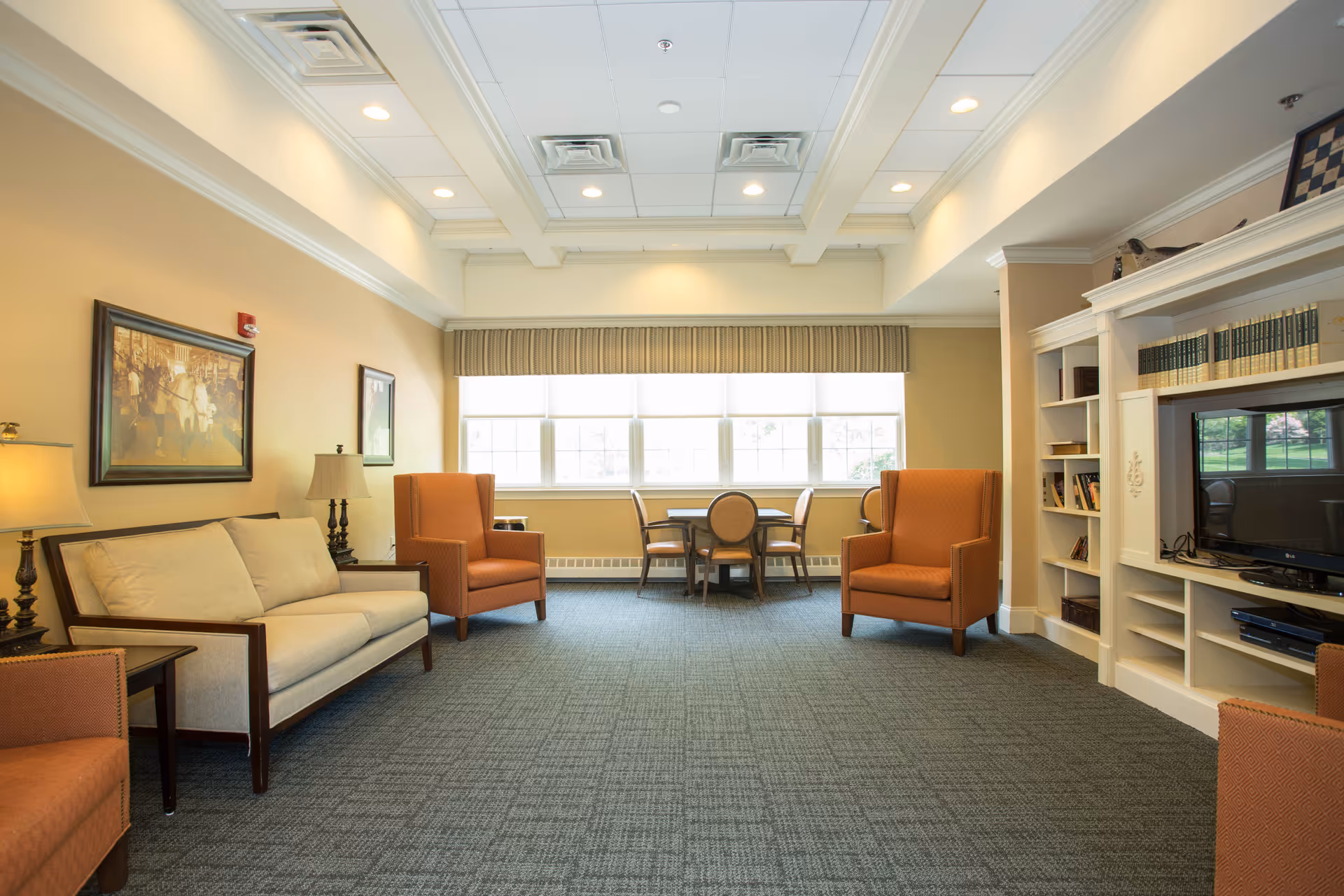 A well-lit living room with beige walls and a large window with blinds. The room features a beige sofa, two orange armchairs, a small round table with four chairs near the window, and built-in white shelves holding books and a flat-screen TV. The ceiling has recessed lighting and decorative molding.