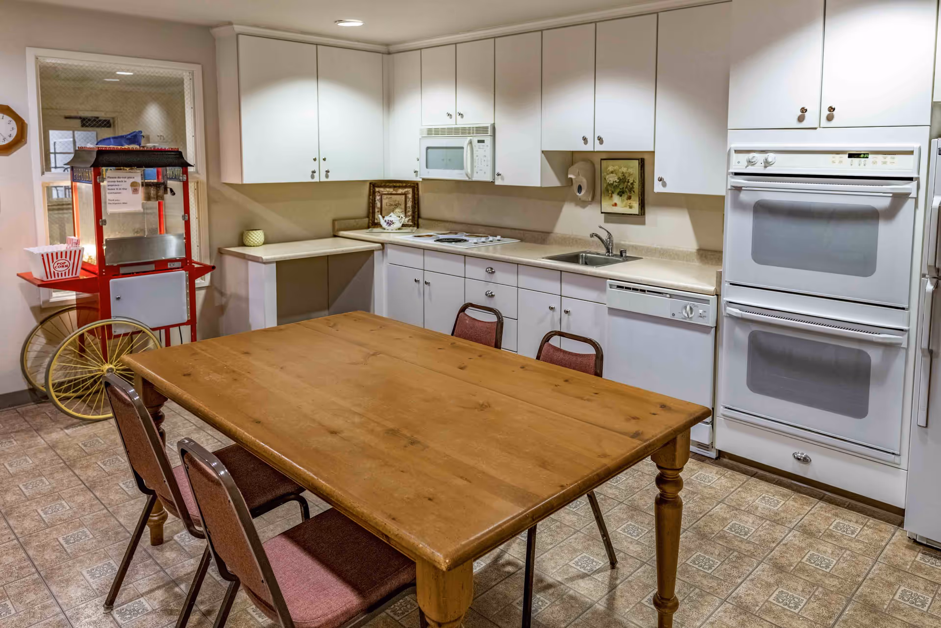 A kitchen area with white cabinets, a microwave, double oven, dishwasher, and a sink. There is a wooden table with four chairs in the center of the room. In the corner, there is a red popcorn machine with a popcorn container on its side shelf. The floor has patterned tiles and the walls are light-colored.