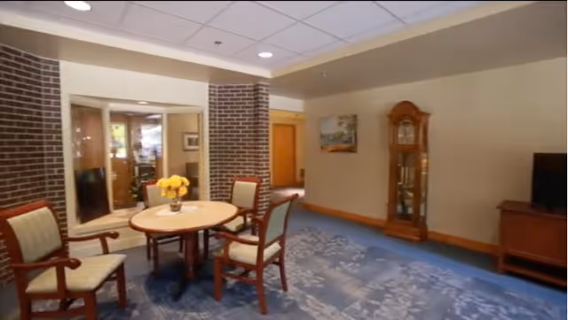 A cozy interior room with a round wooden table surrounded by four cushioned chairs. The table has a vase with yellow flowers. The room features brick pillars, a grandfather clock against the wall, a painting, and a television on a wooden stand. The floor is covered with a blue patterned carpet.