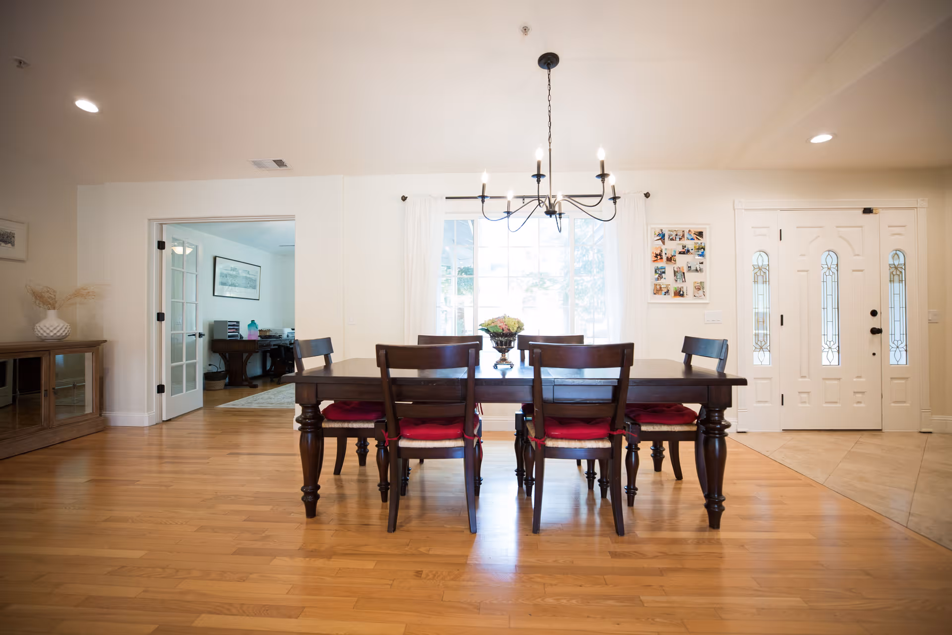 Bright dining room with a dark wood table and six chairs under a chandelier, hardwood floors, and an entry door and window in the background.