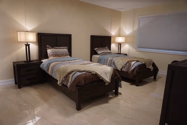 A simple bedroom with two matching twin beds, dark wood nightstands and lamps on a tiled floor.