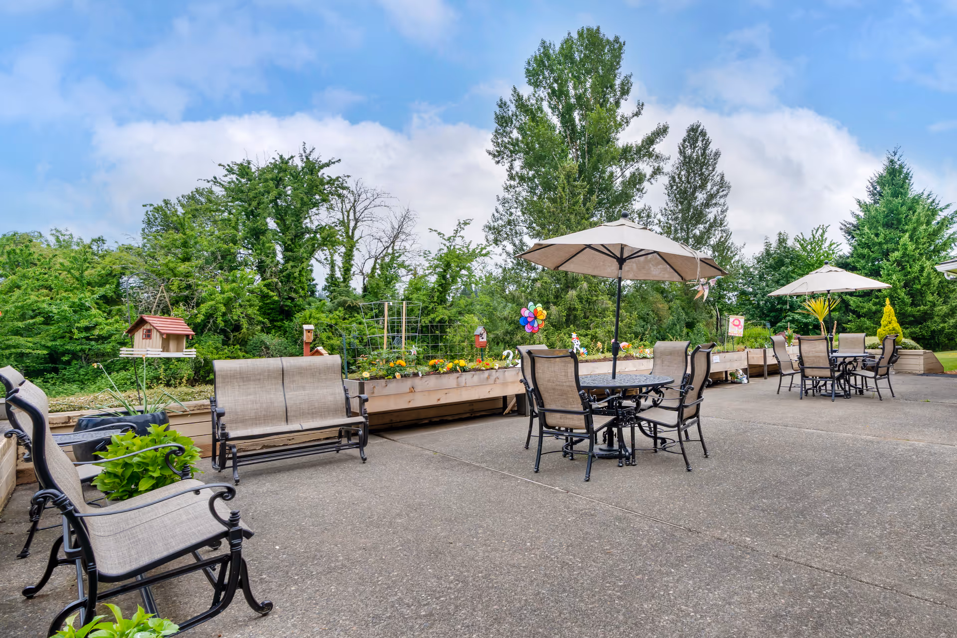 Outdoor patio area with multiple seating arrangements including chairs and benches with beige cushions, two tables with large beige umbrellas, raised garden beds with flowers and plants, colorful garden decorations, and tall green trees in the background under a partly cloudy blue sky.