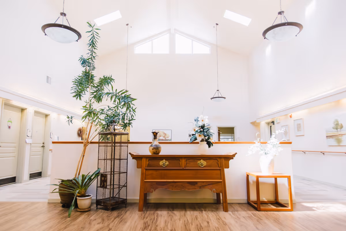 Bright and airy interior hallway of Woodland Heights Assisted Living with high vaulted ceilings and skylights. The space features a wooden sideboard with decorative vases and flowers, potted plants, and framed artwork on the walls. Doors to resident rooms are visible on the left side, and the floor is wood-patterned.