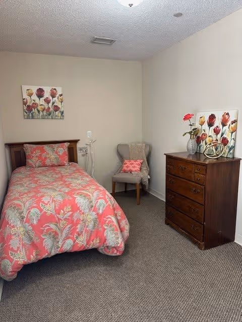 A small bedroom with a single bed covered in a pink floral bedspread. Above the bed is a painting of flowers. To the right of the bed is a wooden dresser with a vase holding a single red flower, a decorative item, and another floral painting on the wall above it. A gray upholstered chair with a pink pillow and a beige throw blanket is positioned in the corner. The room has beige walls and carpeted flooring.