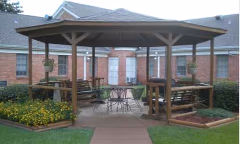 Outdoor gazebo with benches and a metal table under a shaded wooden pavilion, brick building behind.