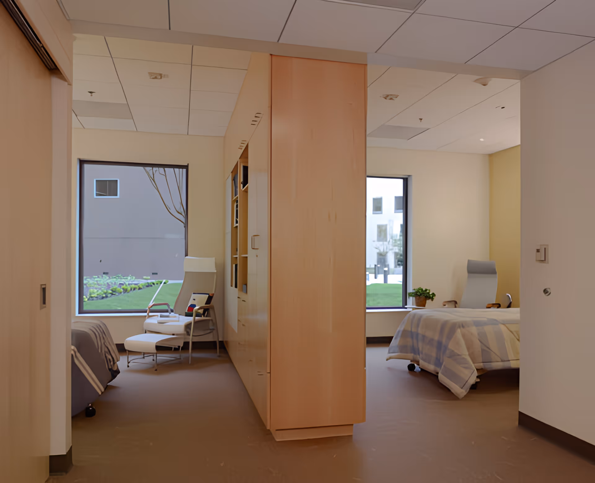 Interior view of a senior living facility room with two separate sleeping areas divided by a wooden partition. Each area has a bed, a comfortable chair, and large windows showing an outdoor garden area.