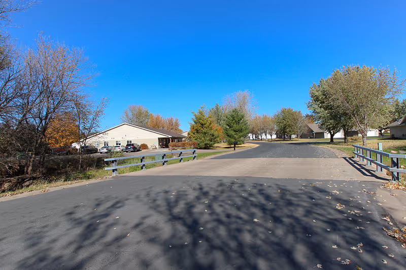 Driveway and entrance road leading to a single-story assisted living building with trees and parked cars under a clear blue sky.