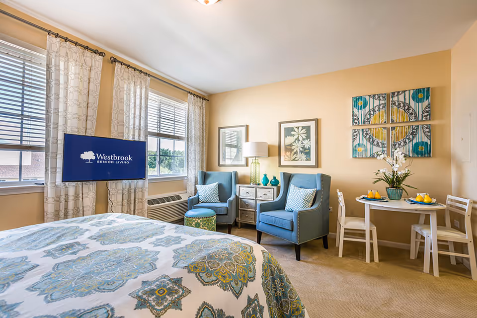 A bright and cozy senior living bedroom with a bed covered in a blue and green patterned bedspread. Two large windows with white patterned curtains let in natural light. A flat-screen TV mounted on the wall displays the Westbrook Senior Living logo. The room features two blue armchairs with patterned cushions, a small round ottoman, a side table with a lamp and decorative vases, and a small dining table with two chairs and a flower arrangement. The walls are decorated with framed artwork.