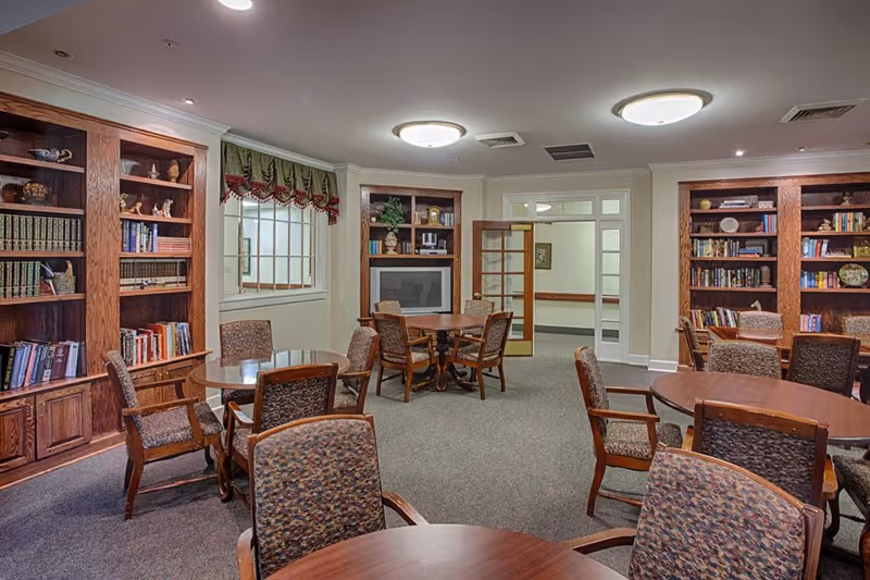 A cozy common room with several round wooden tables and cushioned chairs arranged around them. The room features built-in wooden bookshelves filled with books and decorative items. There is a window with a valance and a glass door leading to another area. The lighting is provided by ceiling fixtures, and the carpet is a neutral color.