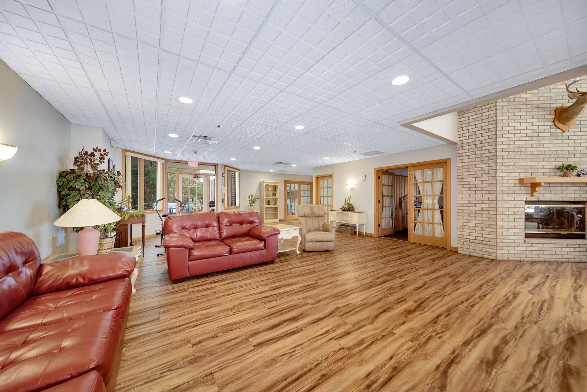 A spacious living room area with wood flooring, two red leather sofas, a beige recliner chair, a white coffee table, and a brick fireplace with a mounted deer head above it. The room has a white tiled ceiling with recessed lighting, large windows, glass doors, and several plants and lamps for decoration.