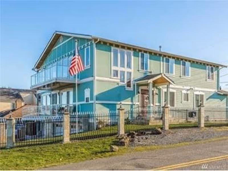 Two-story teal-green residential building with an American flag and a fenced yard seen from the street.