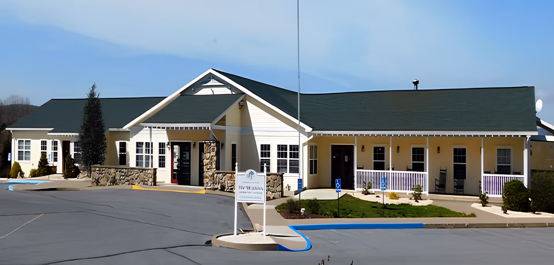 Exterior view of Meadows At Maria Joseph Manor, a single-story building with a green roof and light-colored walls. The entrance features stone accents and a covered porch area with a sign in front. There are several windows and a small landscaped area with bushes and a wheelchair accessible parking space.