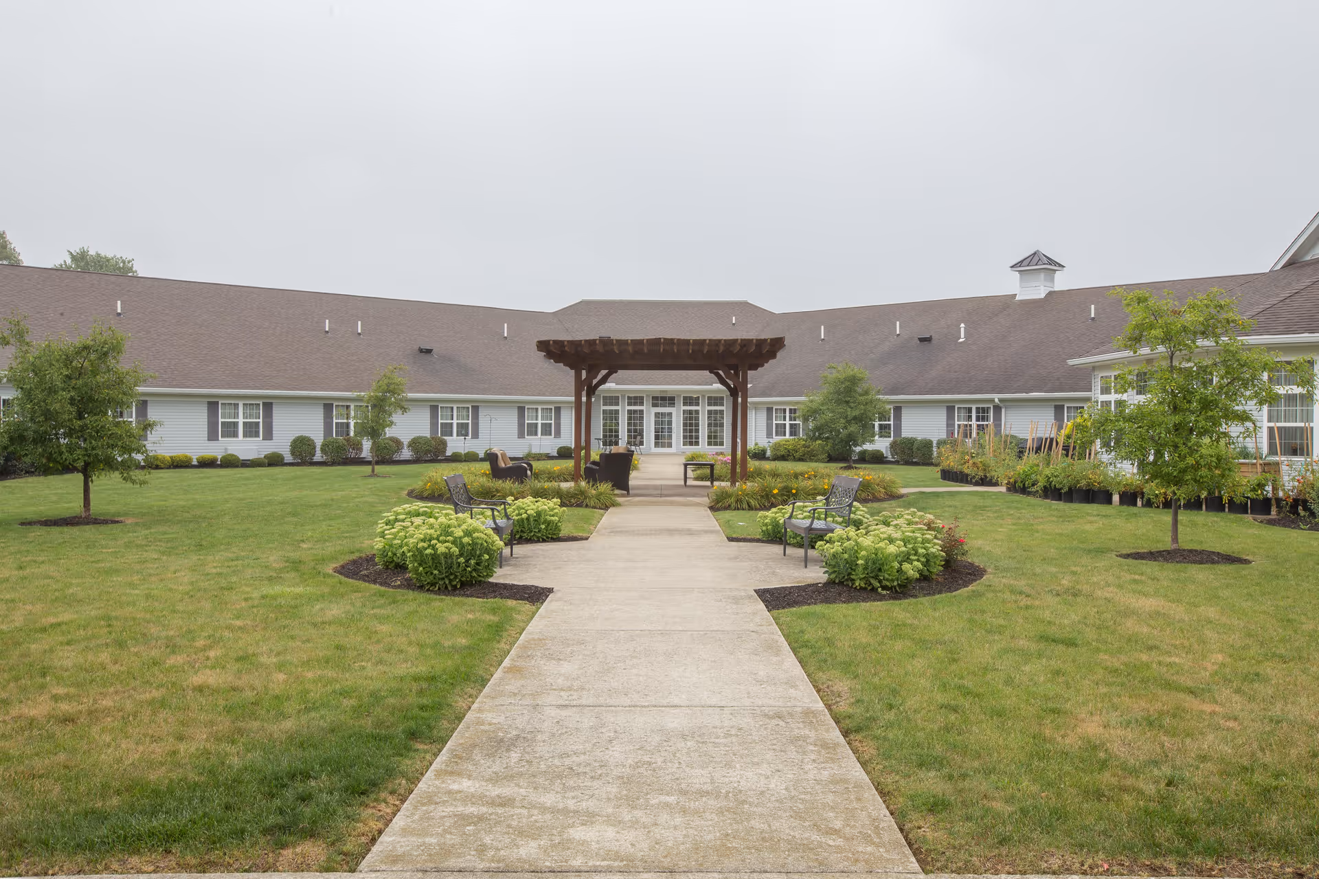 A courtyard area of a senior living facility with a concrete walkway leading to a wooden pergola. The courtyard is surrounded by a single-story building with multiple windows and a brown shingled roof. There are green lawns, small trees, shrubs, and benches along the walkway.