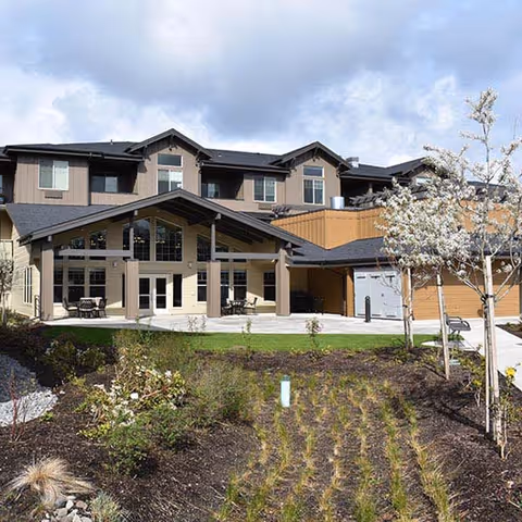 Exterior view of a two-story senior living facility building with a covered entrance, large windows, and landscaped garden with young trees and plants in front.