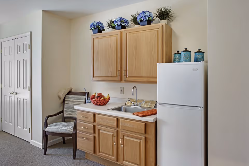 A small kitchenette area with wooden cabinets above and below a countertop with a sink. On the countertop is a red fruit bowl with various fruits and an orange towel. To the right is a white refrigerator with three blue decorative canisters on top. To the left of the kitchenette is a cushioned wooden chair with a hat hanging on the backrest. The walls are light-colored and there is a double-door closet on the far left.