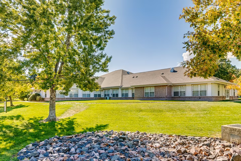 Exterior view of a single-story senior living facility building with a sloped roof, surrounded by green grass, trees, and a rock bed in the foreground under a clear blue sky.