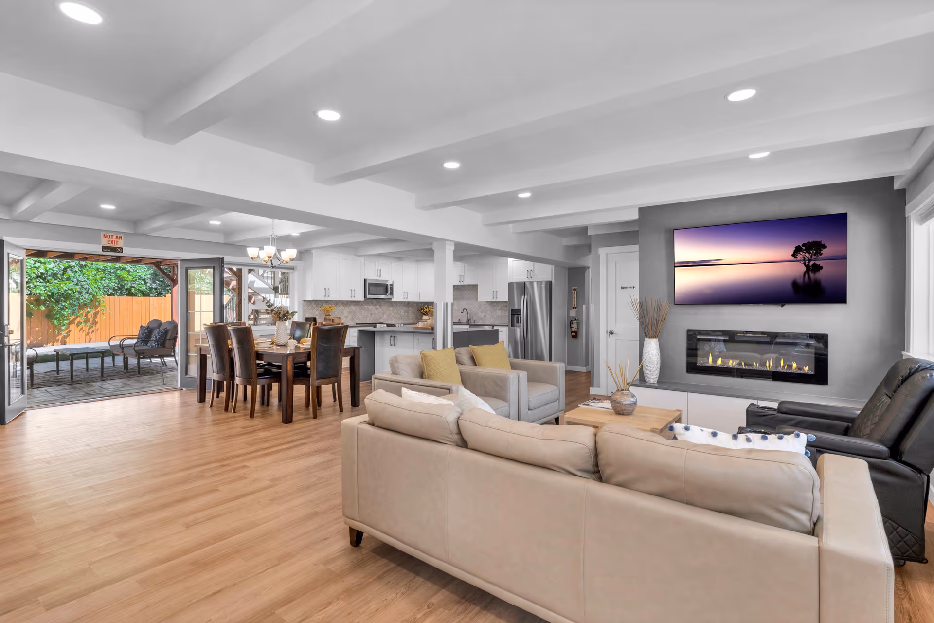 A spacious, modern living area with beige sofas, two armchairs, and a black recliner arranged around a wooden coffee table. A flat-screen TV displaying a serene landscape is mounted above a contemporary fireplace on a gray accent wall. The open floor plan includes a dining area with a wooden table and six chairs, and a kitchen with white cabinets, stainless steel appliances, and a tiled backsplash. Large glass doors open to an outdoor patio with seating and greenery.