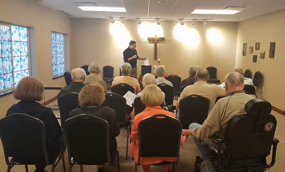 A group of elderly people seated in rows of chairs facing a man standing at the front of a room with a wooden cross on the wall behind him. The room has beige walls, stained glass windows on the left, and several small framed pictures on the right wall. The man appears to be speaking or reading from a paper.