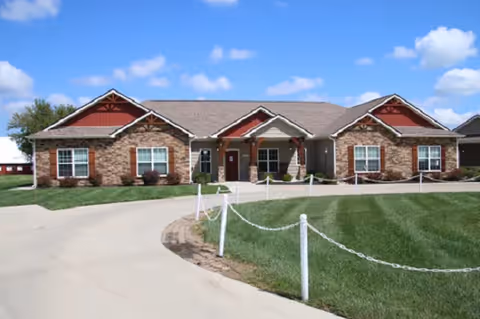Single-story brick-and-stone senior living building with a central covered entrance, manicured lawn, and paved driveway under a blue sky.
