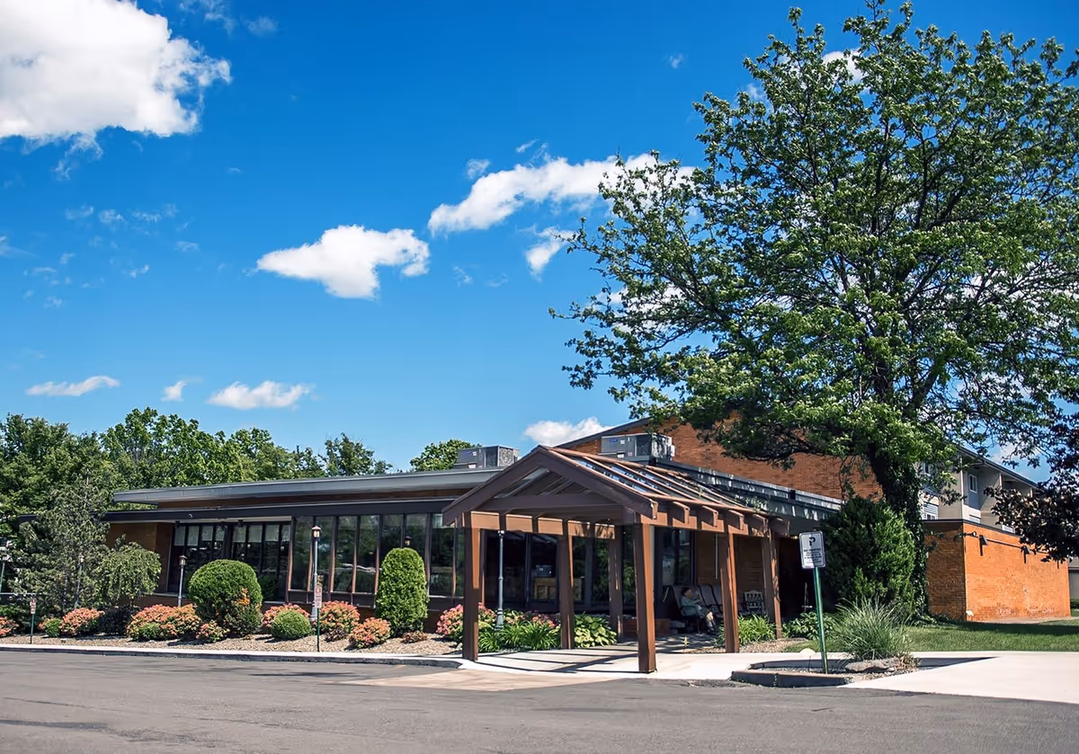 Exterior view of The Commons on St. Anthony showing a brick building with glass frontage, a wooden entrance canopy, and landscaped shrubs.