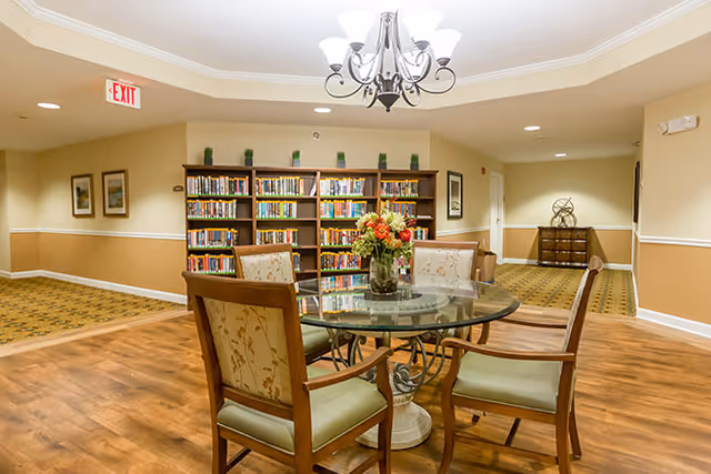A cozy interior common area with a round glass table surrounded by four wooden chairs with cushioned seats and backs. A vase with a colorful flower arrangement is placed on the table. Behind the table is a bookshelf filled with books and decorative plants on top. The room has warm beige walls with white trim, framed artwork, and a chandelier hanging from the ceiling. The floor is a combination of wood and patterned carpet.