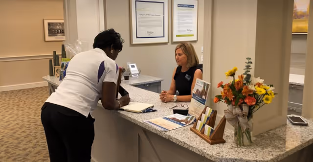 A receptionist sits behind a granite front desk speaking with a visitor who is signing paperwork in a senior living facility lobby.