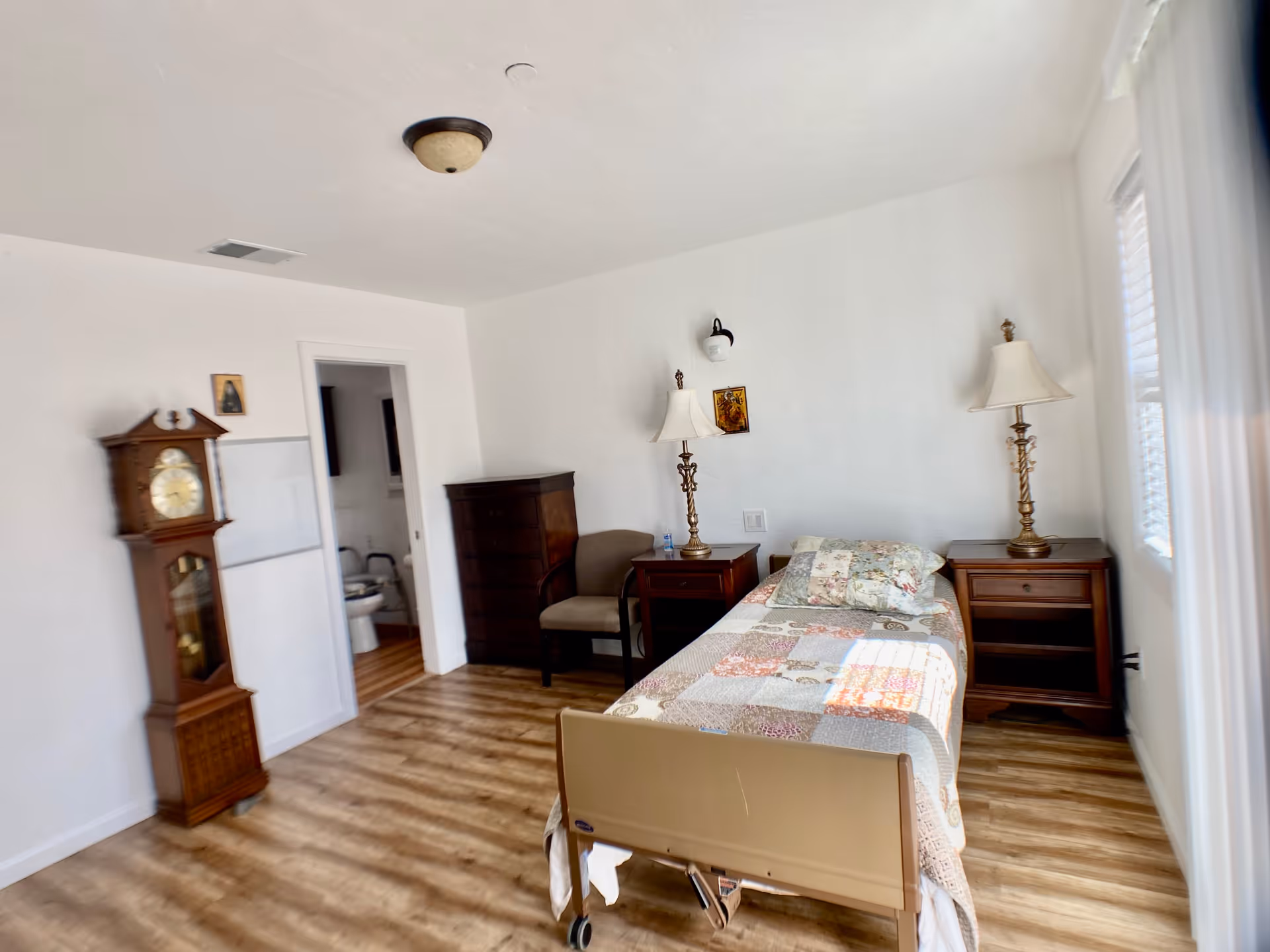 A simple elderly care bedroom with a single bed covered in a patchwork quilt, two wooden nightstands with matching lamps, a wooden dresser, a chair, and a grandfather clock. There is a doorway leading to a bathroom with a toilet visible. The room has wood flooring and white walls with a window letting in natural light.