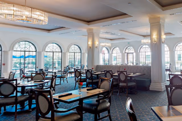 Spacious dining room with multiple tables and chairs arranged neatly. Large arched windows allow natural light to fill the room. Elegant chandeliers and wall sconces provide additional lighting. The room has a carpeted floor and a mix of booth and chair seating.