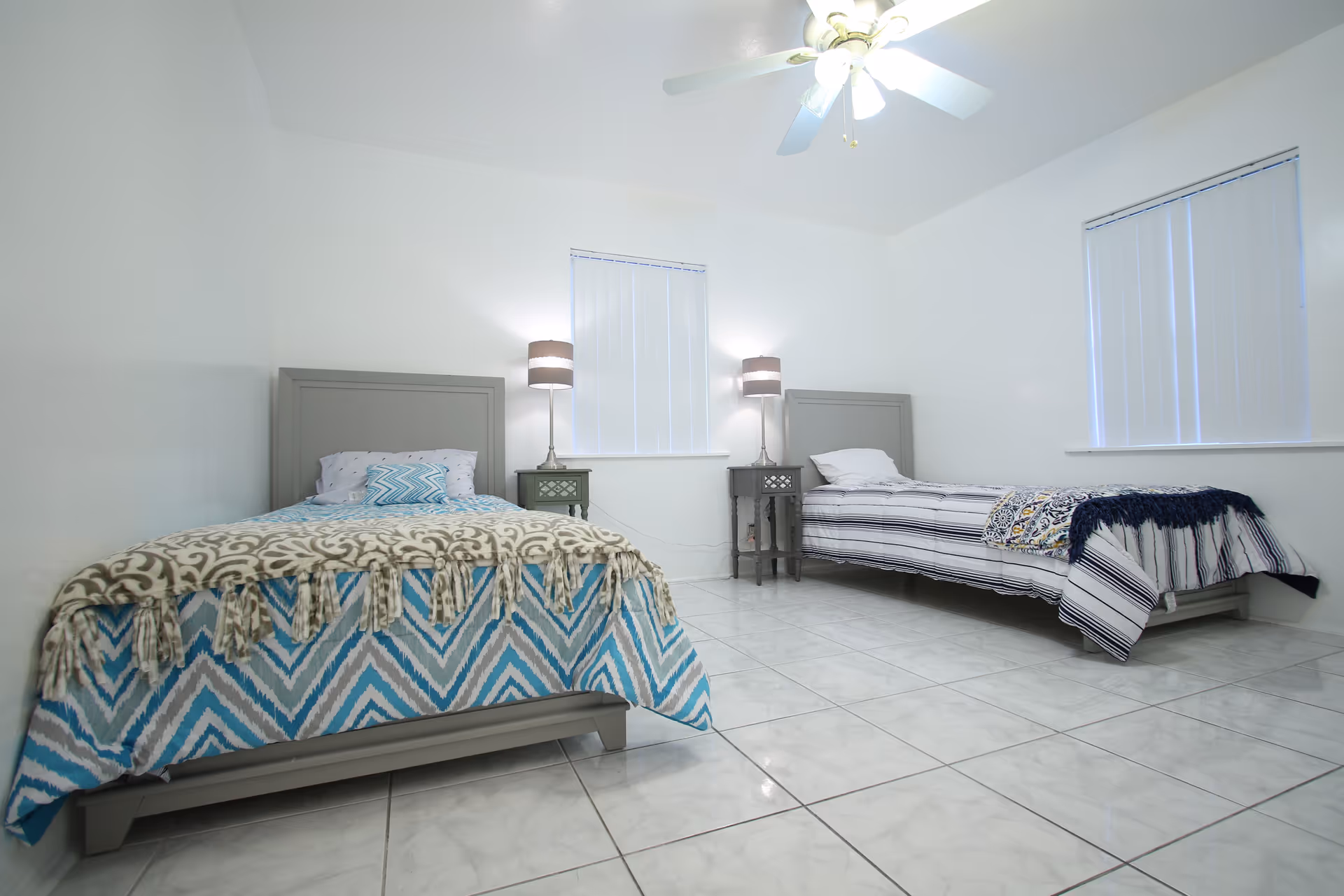 A bright bedroom with two single beds, each with patterned bedding and a pillow. Between the beds are two small nightstands with matching lamps. The room has white walls, two windows with vertical blinds, and a ceiling fan with lights. The floor is tiled with white marble-like tiles.