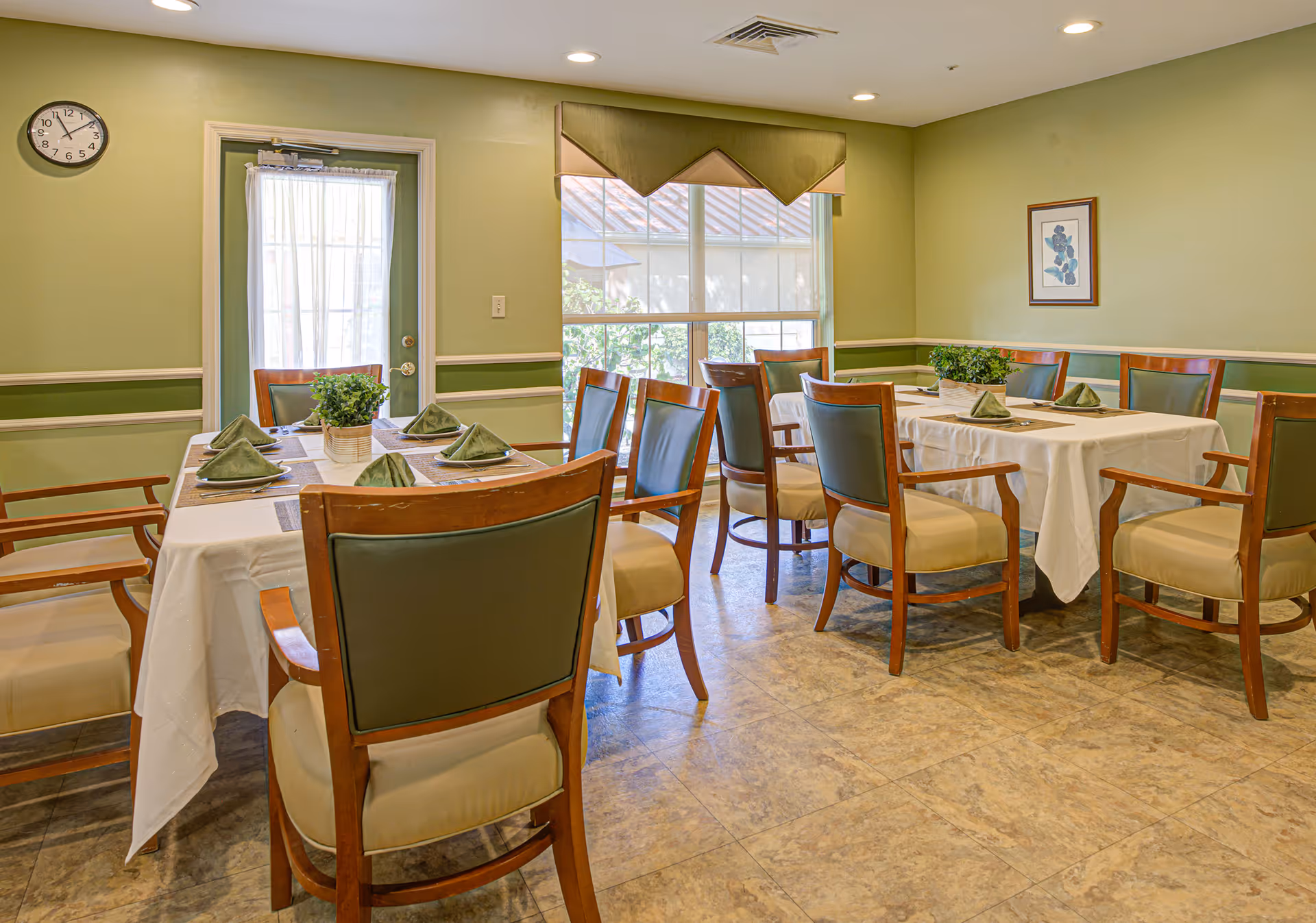 A bright dining room with multiple tables set with white tablecloths, green napkins, wooden chairs, and potted plants.