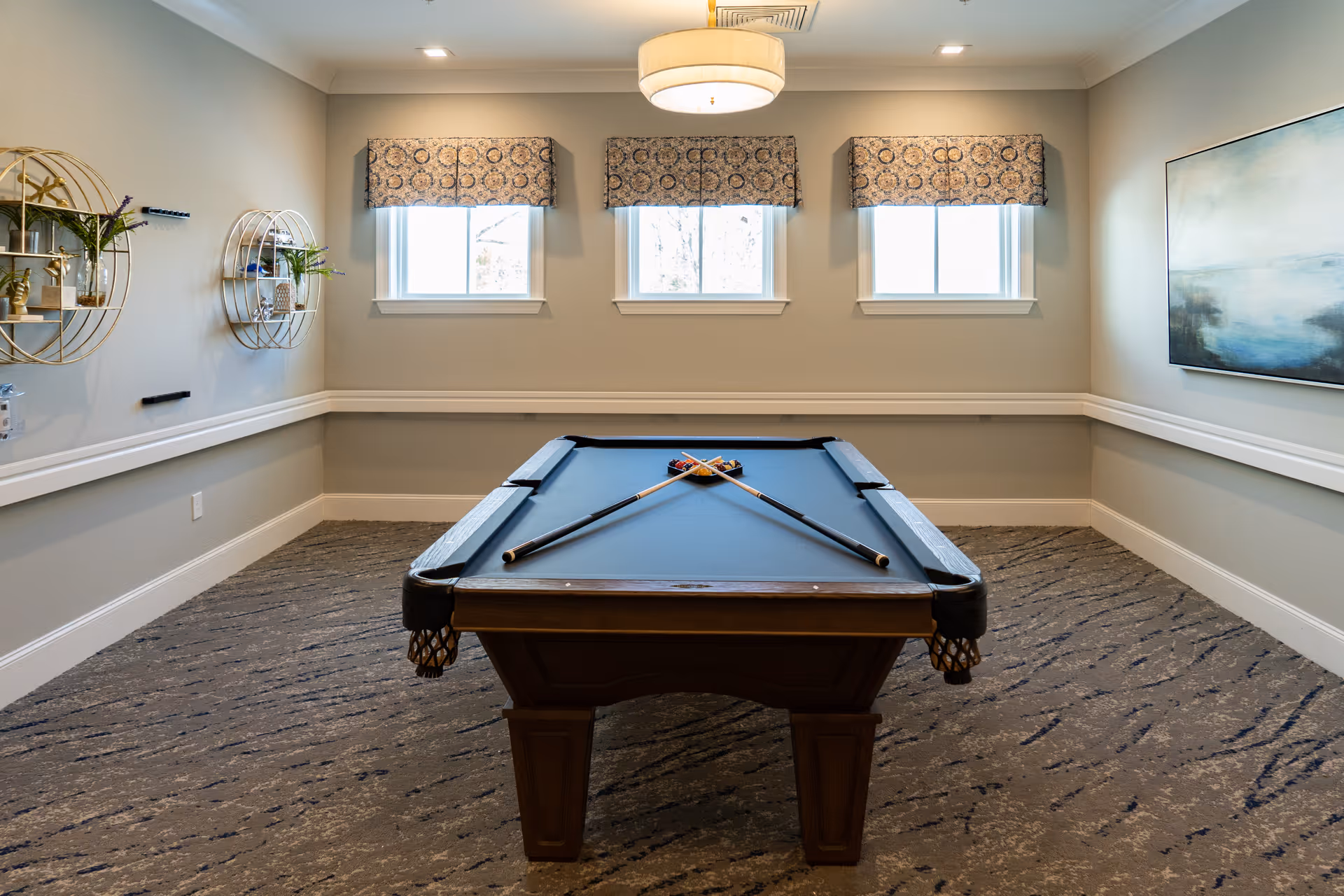 A well-lit room with a pool table in the center, two pool cues crossed on the table, three windows with patterned valances, a modern ceiling light fixture, decorative wall shelves with plants on the left wall, and a large abstract painting on the right wall.