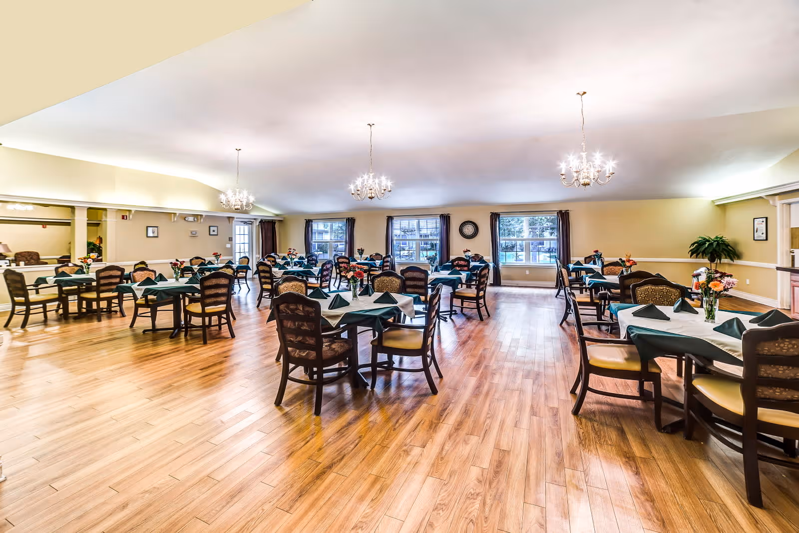 A spacious dining room with multiple tables covered with white tablecloths and green napkins. Each table is surrounded by wooden chairs with cushioned seats. The room has wooden flooring, three chandeliers hanging from the ceiling, and large windows with dark curtains allowing natural light to enter. There are flower arrangements on each table and a clock on the far wall.
