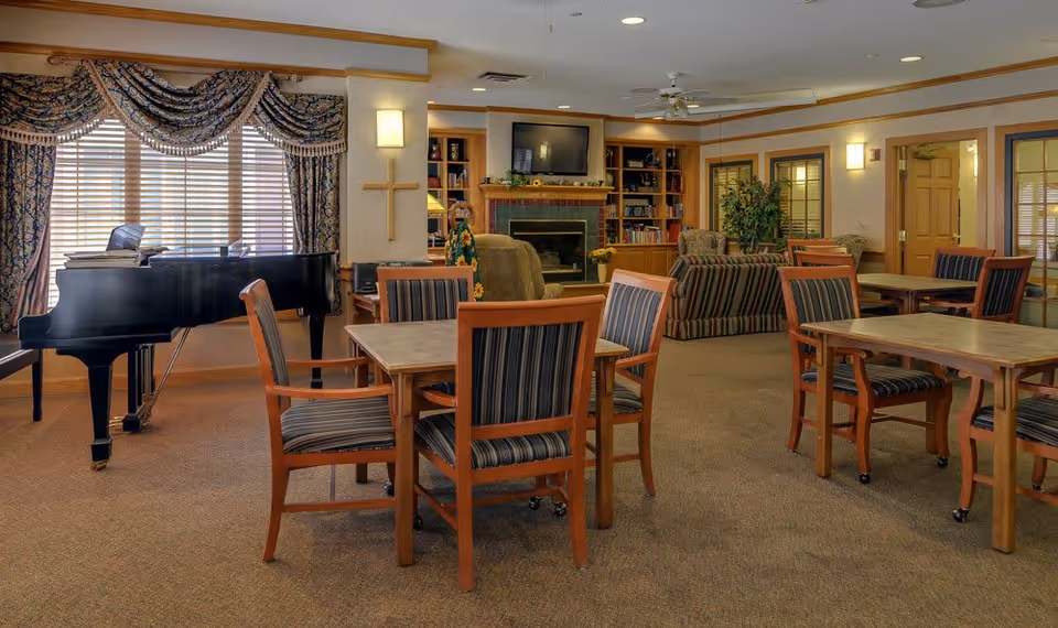 A cozy senior living common area with multiple wooden tables and striped cushioned chairs. A black grand piano is positioned near a window with patterned curtains. In the background, there is a fireplace with a TV mounted above it, built-in bookshelves, and comfortable seating including armchairs and a sofa. The room is warmly lit with wall sconces and ceiling lights.