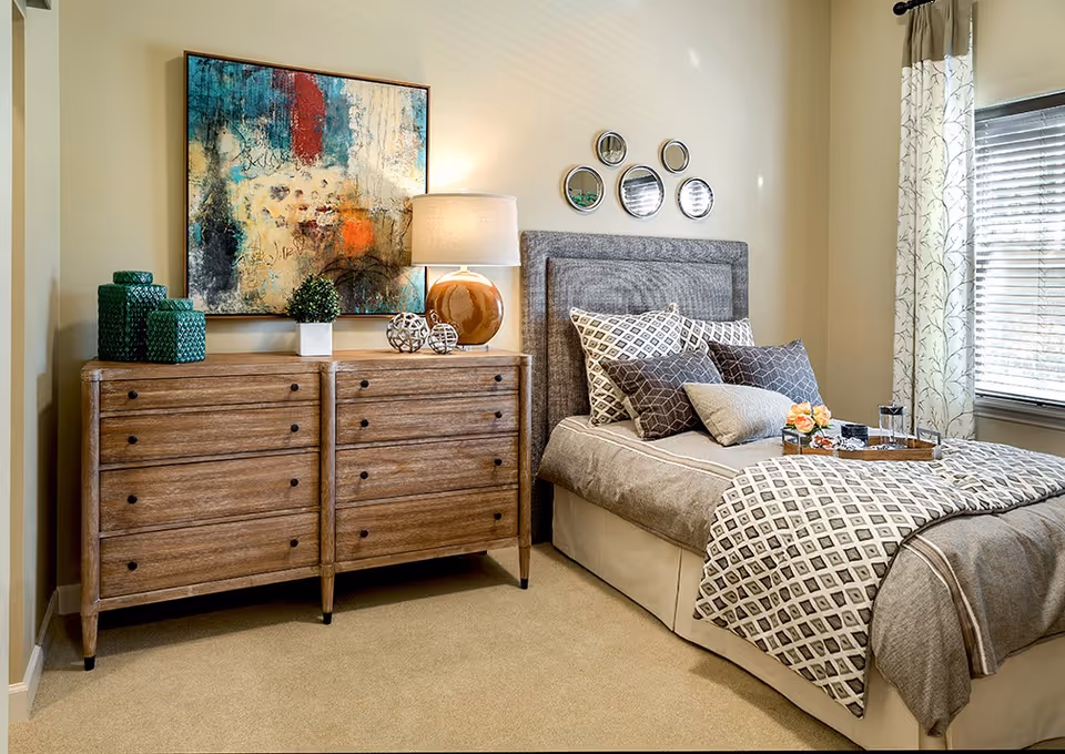 Cozy bedroom with a gray upholstered bed, patterned pillows and coverlet, a wooden double dresser topped with a lamp and abstract artwork, and a window with curtains.