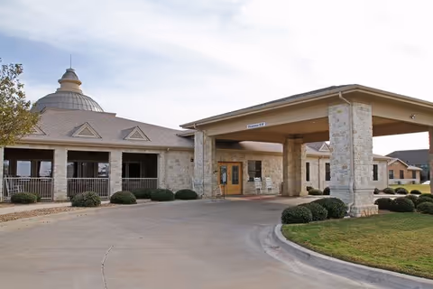 Stone-faced senior living facility entrance with a covered porte-cochere, circular driveway, and a dome-topped roof.