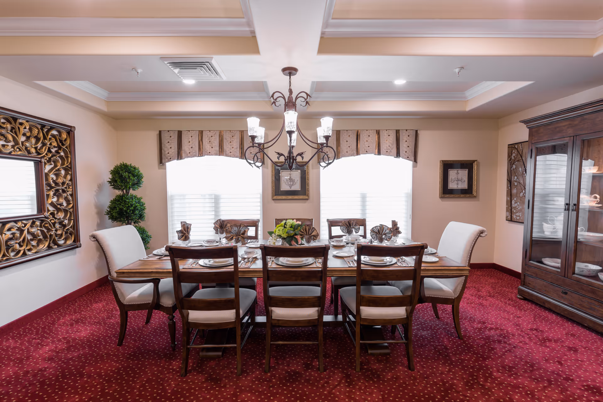 A formal dining room with a wooden dining table set for eight people, featuring folded napkins, plates, and silverware. The room has a red carpet with a dotted pattern, two large windows with valances, a chandelier hanging above the table, a decorative mirror on the left wall, a potted plant, framed artwork, and a wooden china cabinet on the right side displaying dishes.