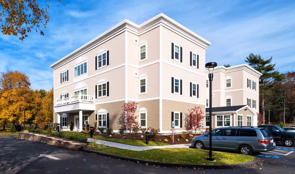 Exterior view of a three-story senior living facility building with beige and white siding, multiple windows with black shutters, a small balcony, and a parking lot with cars. Trees with autumn foliage surround the building under a clear blue sky.