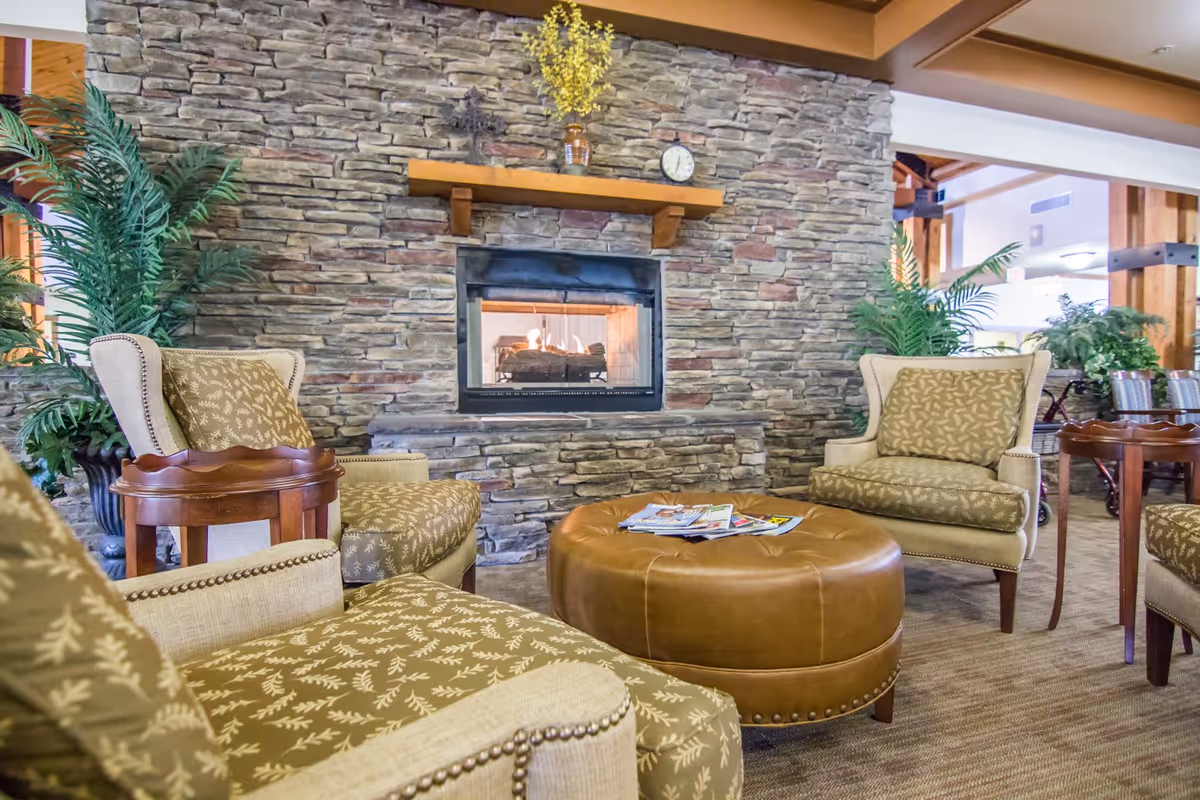 A cozy living room area with a stone fireplace in the center. The fireplace has a wooden mantel with a small clock and a decorative vase with yellow flowers. Surrounding the fireplace are four upholstered armchairs with patterned cushions and a round brown leather ottoman in the middle, holding some magazines. There are green potted plants on either side of the fireplace, and wooden beams and furniture are visible in the background.