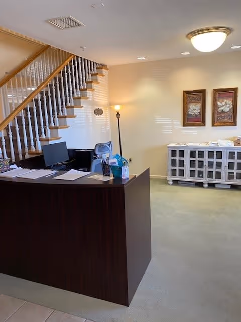 Reception area with a dark wooden desk holding papers, a computer monitor, and office supplies. Behind the desk is a staircase with wooden steps and white balusters. To the right, there is a white cabinet with glass-paneled doors and two framed floral artworks hanging above it. The room is softly lit with a floor lamp and a ceiling light fixture.