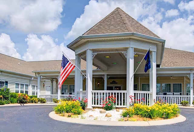 Front entrance of a senior living facility with a covered circular driveway, white railings, and two flagpoles displaying the American flag and another flag. The building has beige siding and a shingled roof, with flower beds featuring yellow and red flowers in front of the entrance.