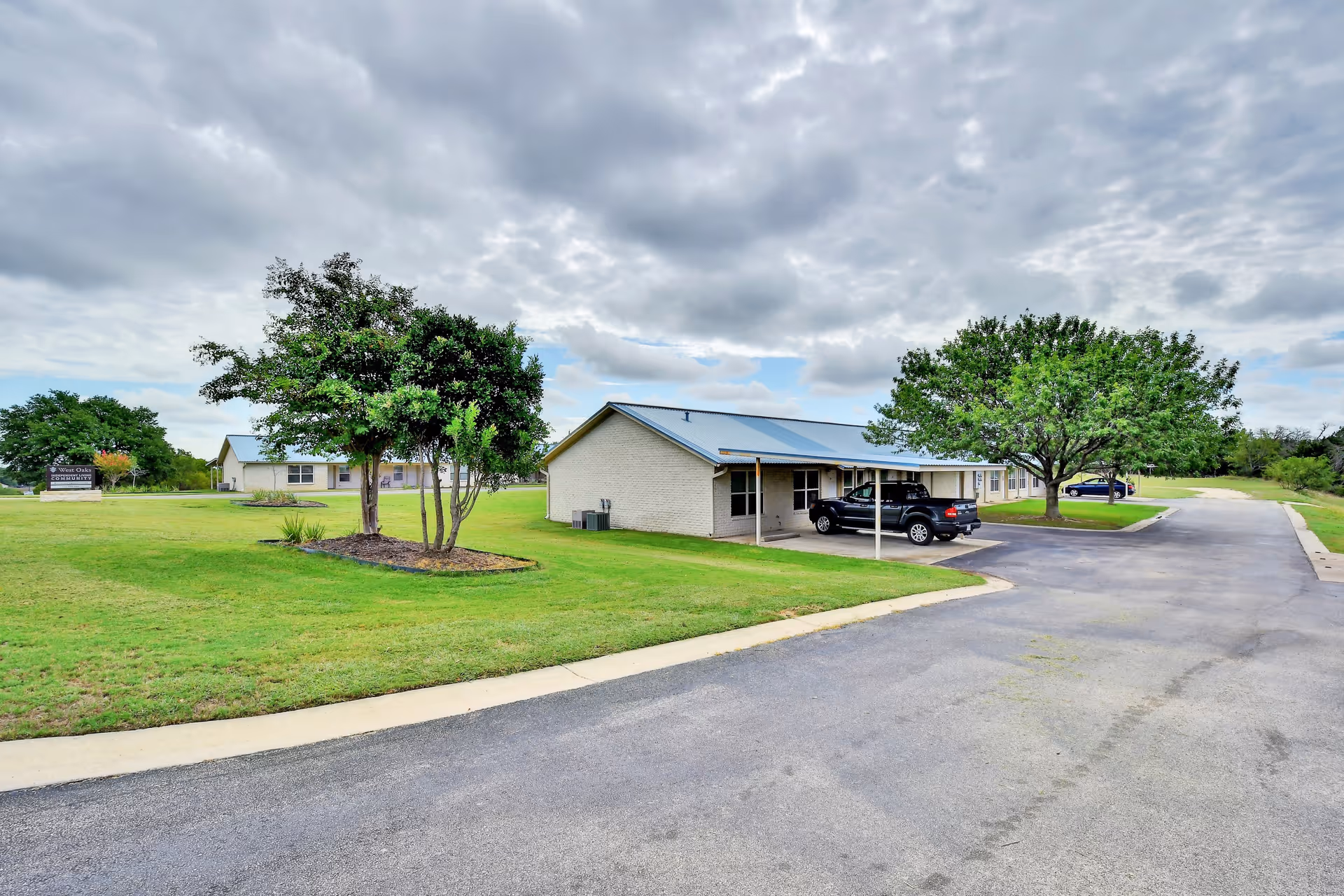 Single-story senior living buildings with carports, parked cars, green lawns and trees under a cloudy sky.